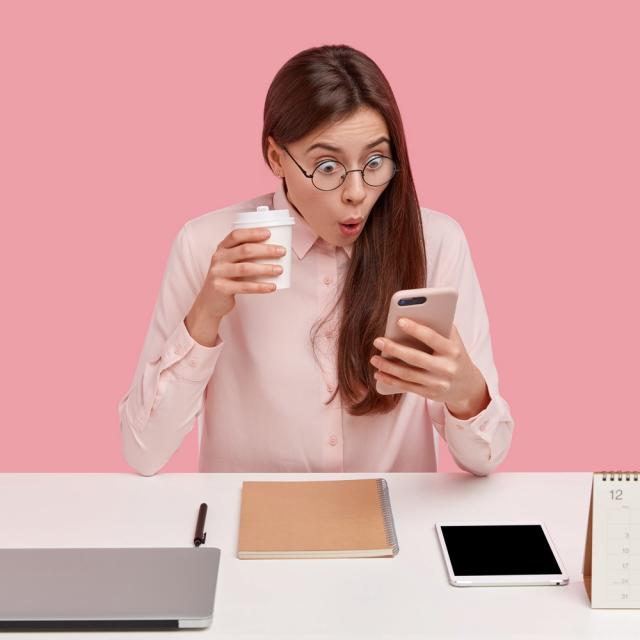 Photo of attractive young woman reads shocking news on mobile phone, watches video in social networks, drinks coffee from disposable cup, being office perfectionist, uses electronic gadgets.