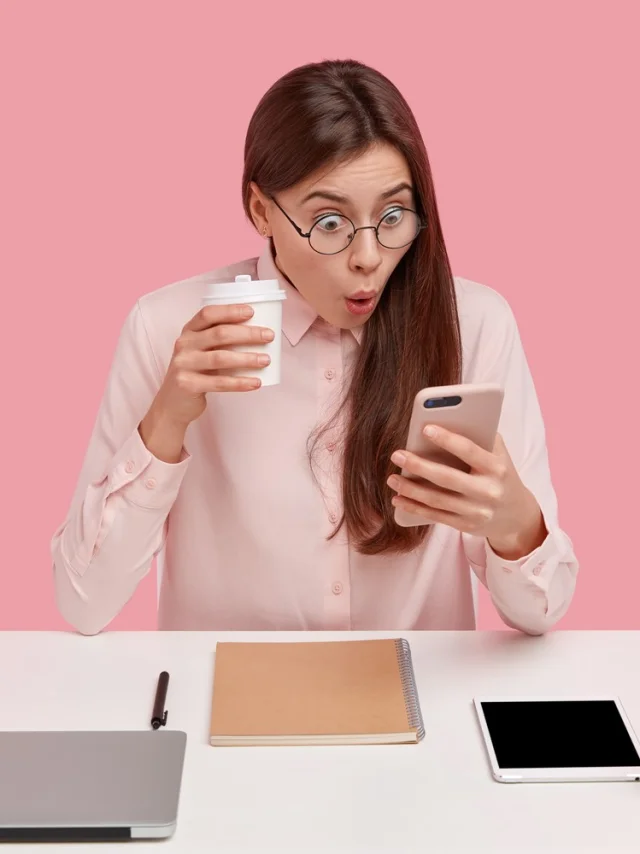 Photo of attractive young woman reads shocking news on mobile phone, watches video in social networks, drinks coffee from disposable cup, being office perfectionist, uses electronic gadgets.